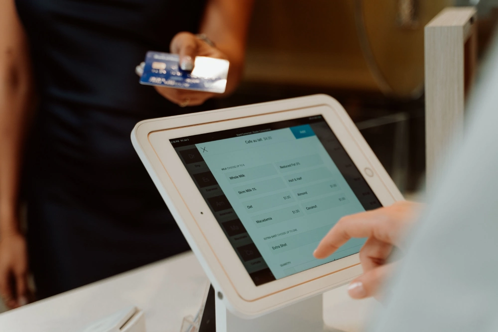 A cashier using a white touchscreen POS terminal to select order details while a customer hands over a credit card, demonstrating integrated hardware, software, and payments.